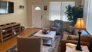 Ein Wohnzimmer mit Fernseher, Couchtisch, Sofa, Sessel und Lampe. Eine Tür und ein Fenster mit Jalousien sind vorhanden.