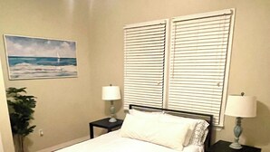 Ein ordentlich bezogenes Bett in einem Zimmer mit großem Fenster, zwei Nachttische mit Lampen, ein Topfpflanze und ein Bild an der Wand.