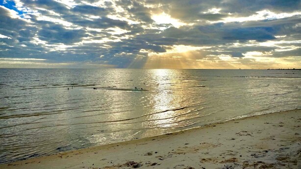 Ein Strand mit breitem Sandufer und ruhigem Wasser, das das Sonnenlicht spiegelt.