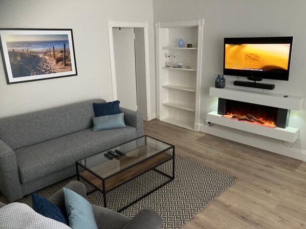 A modern living room with a grey sofa, a glass coffee table, a fireplace, and a flat-screen TV on a white shelf.
