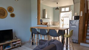 Un salon avec un téléviseur, des livres et un canapé. On y trouve également un coin repas avec des chaises et une table. Une cuisine est visible au fond.