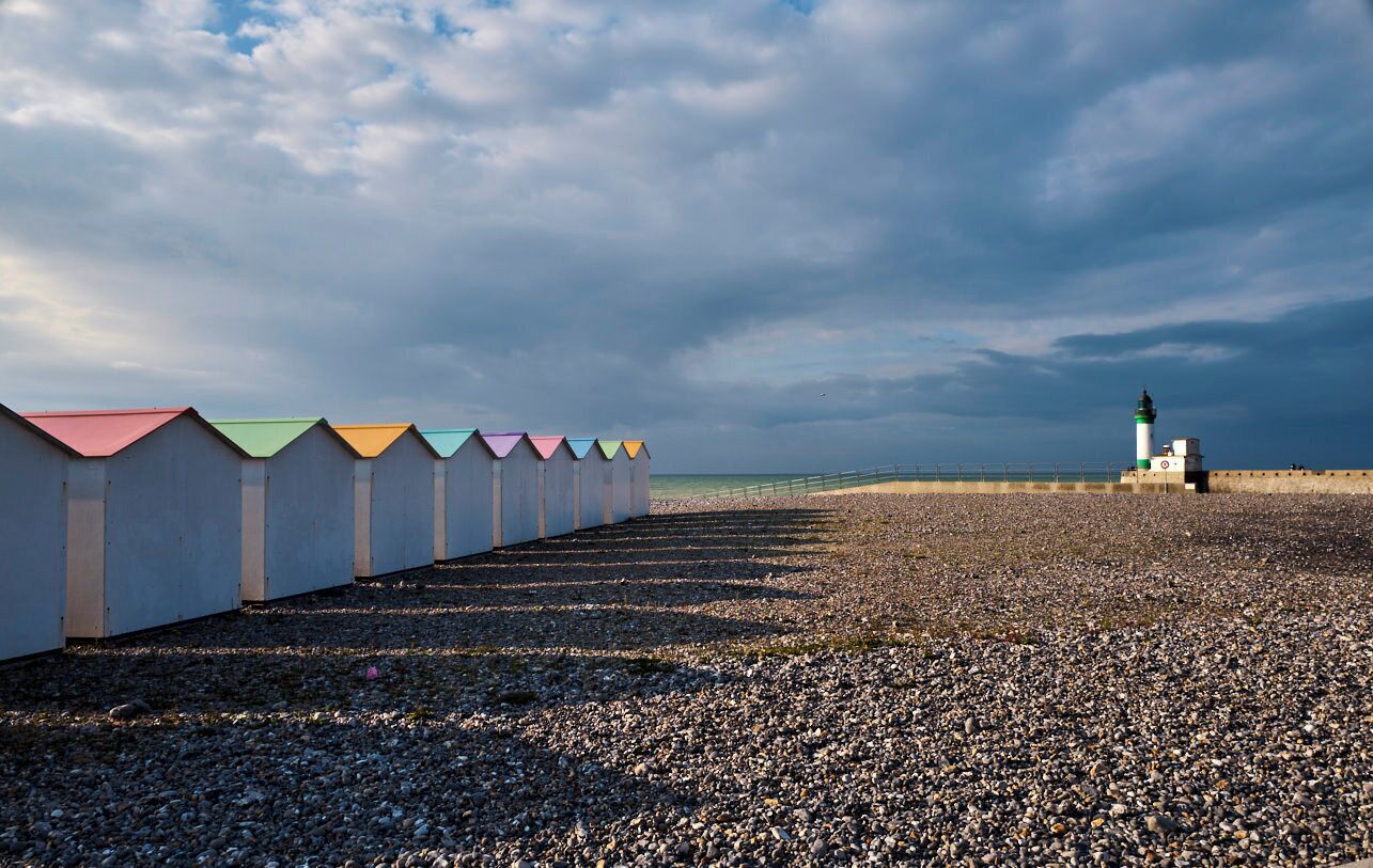 A row of colorful beach huts along a pebbled beach with a lighthouse in the distance under a cloudy sky.