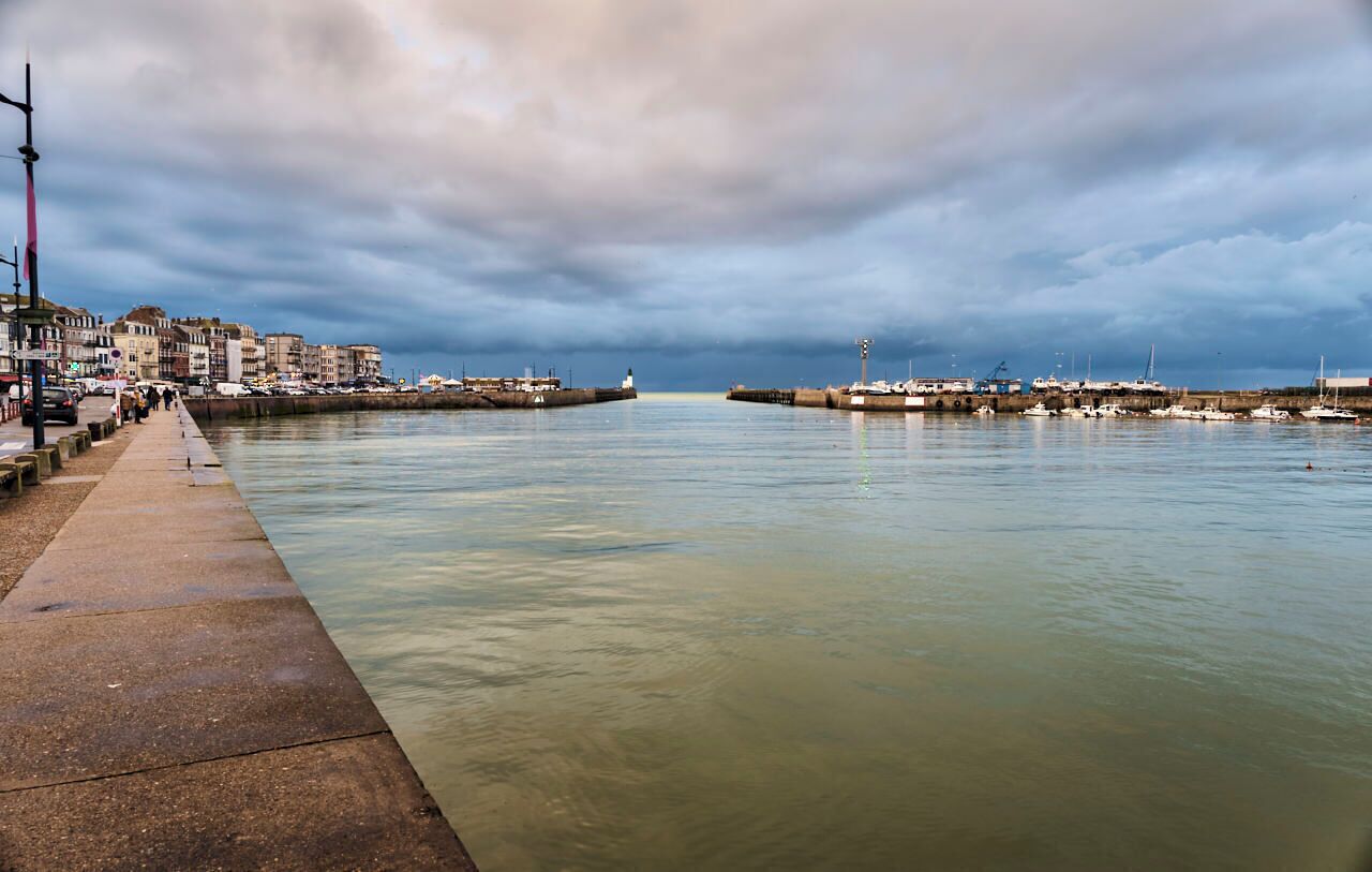 A waterfront promenade with a concrete walkway, a lighthouse in the distance, and a marina with boats.