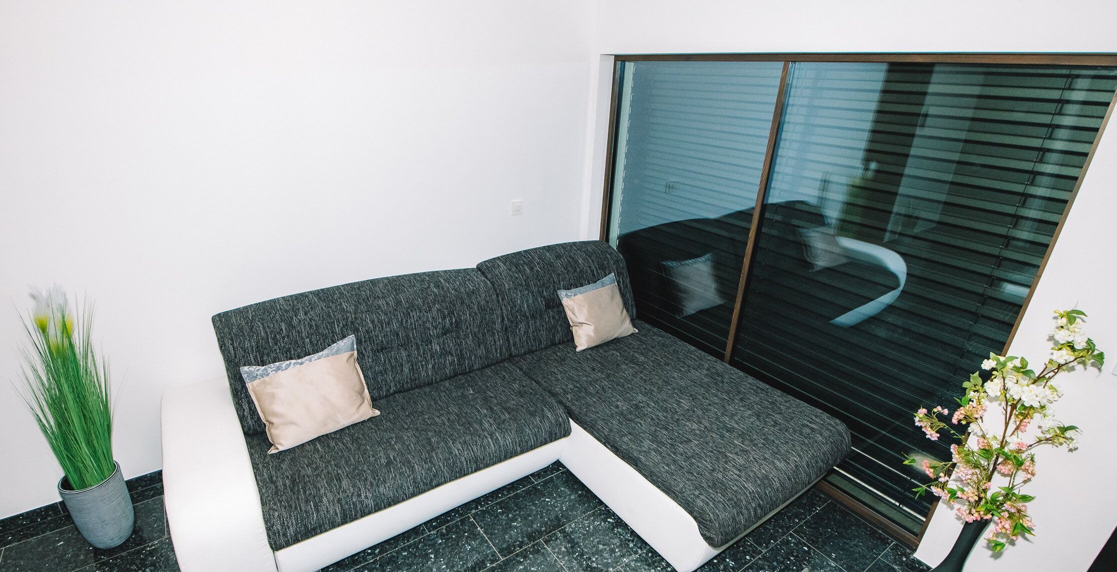 A modern living room with a grey sofa, two cushions, a small potted plant, and a window with blinds.