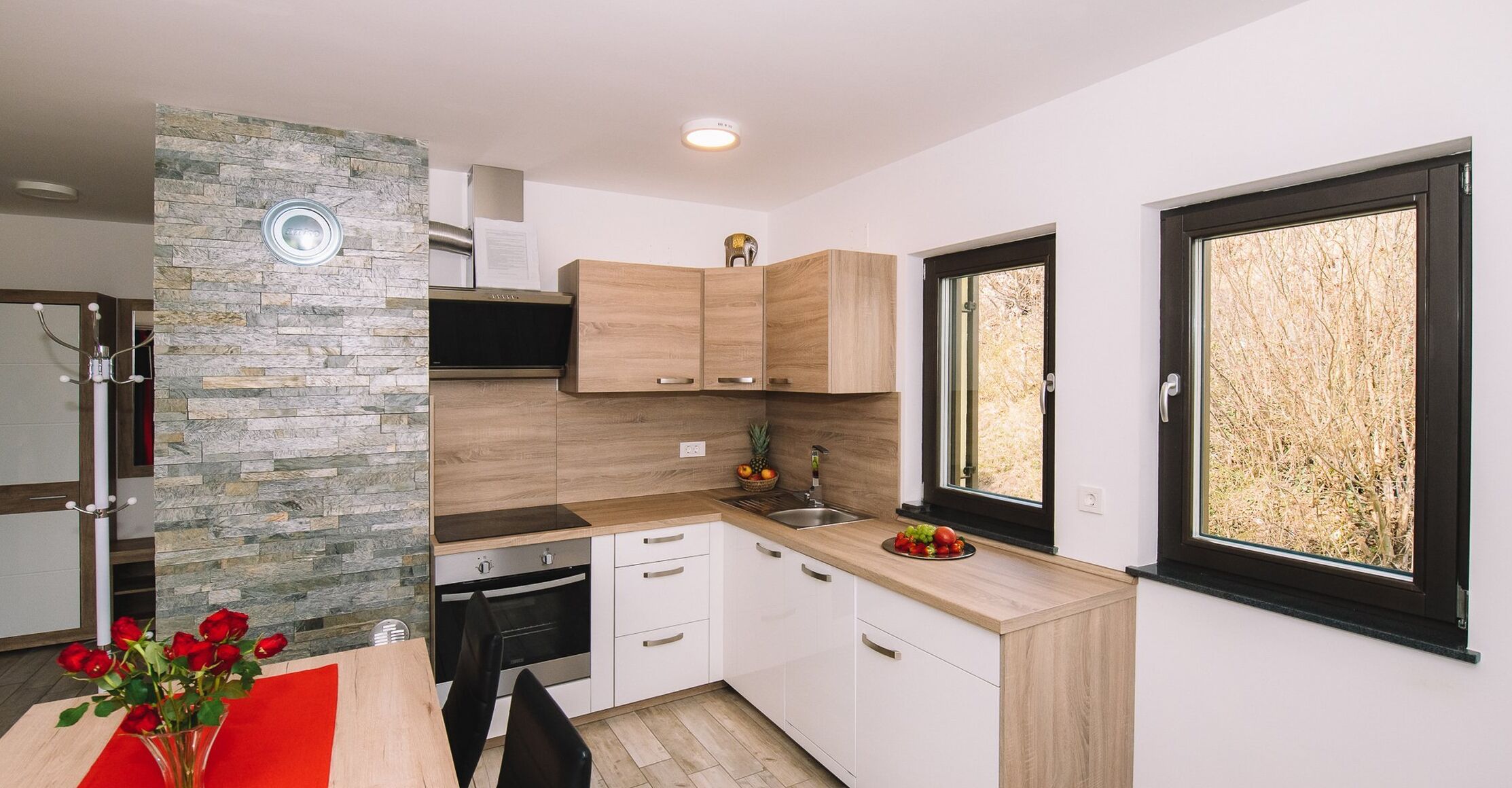 A modern kitchen with a stone accent wall, wooden cabinets, and a view of trees through large windows.