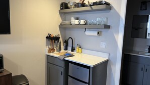 Une cuisine compacte avec un plan de travail blanc, un évier encastré et un petit réfrigérateur. Des étagères au-dessus du plan de travail servent à ranger les ustensiles de cuisine et quelques bouteilles.