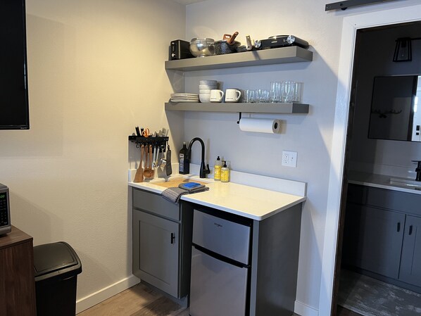 A compact kitchen with a white countertop, a built-in sink, and a small refrigerator. There are shelves above the countertop holding kitchenware and a few bottles. - The Bunkhouse Farm Stay near Madison River! (Three Forks)