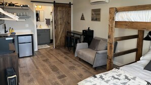 Une pièce compacte comprenant un coin cuisine, un lit recouvert d’une couverture à motifs, un petit canapé et un bureau avec une chaise.