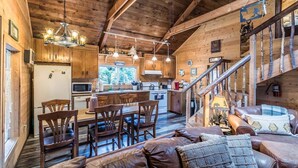 Interior acolhedor de cabana, com área de jantar, cozinha e escada que leva a um quarto no mezanino.