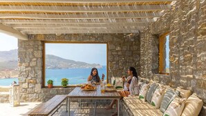 Una habitación con paredes de piedra y un gran ventanal con vistas al mar, una mesa de madera con platos y vasos, y un sofá con cojines.