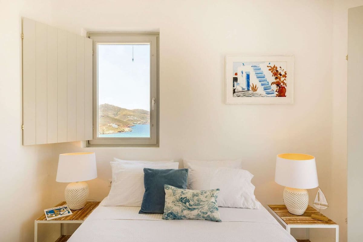 Un dormitorio con cama, dos mesitas de noche con lámparas, una ventana con vistas a un paisaje costero y un cuadro en la pared.