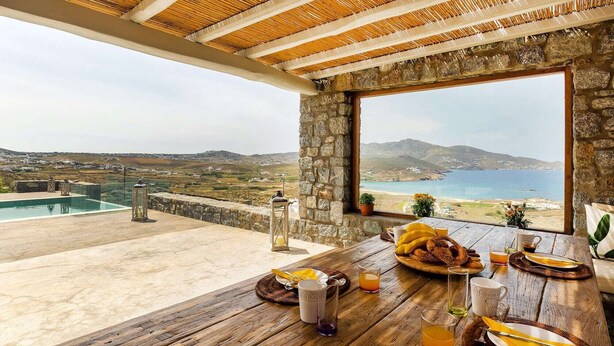 Comedor con vista al mar y colinas, pared de piedra y mesa de madera dispuesta para el desayuno.