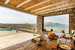 Comedor con vista al mar y colinas, pared de piedra y mesa de madera dispuesta para el desayuno.