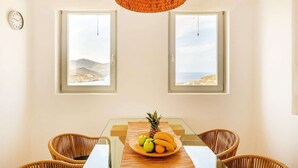 Comedor con una mesa dispuesta para dos, dos sillas de mimbre, una lámpara colgante tejida y dos ventanas con vistas a un paisaje costero.