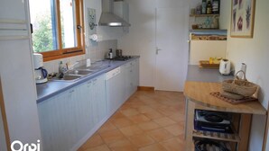 A compact kitchen with a window, a sink, a stove, and a small dining area with a table and shelves. - Maison 2 Chambres Proche Commerces à Louer Pour les Vacances (ARZON)