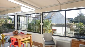 Une terrasse ensoleillée avec une table en bois et des chaises colorées, de grandes fenêtres donnant sur un jardin et un bâtiment.