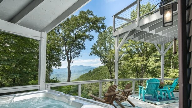 A hot tub on a wooden deck with chairs, overlooking a forested area and mountains.