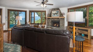 A cozy living room with a stone fireplace, leather sofa, and large windows offering a view of the outdoors.