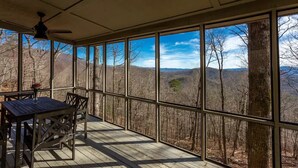 Uma área externa coberta com mesa e cadeiras, grandes janelas com vista para uma paisagem arborizada.