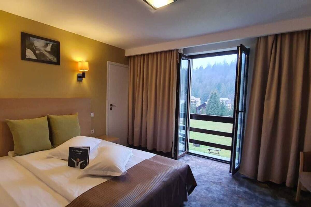 A hotel room with a bed, a bedside table, a nightstand, a window with curtains, and a view of a balcony.
