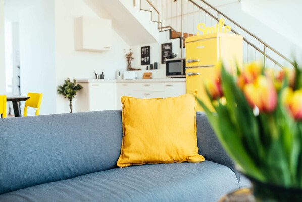 A modern living room with a blue sofa and a mustard yellow pillow, a kitchen in the background, and a staircase leading to an upper level. - Fantastic Loft with Balcony (Berlin)