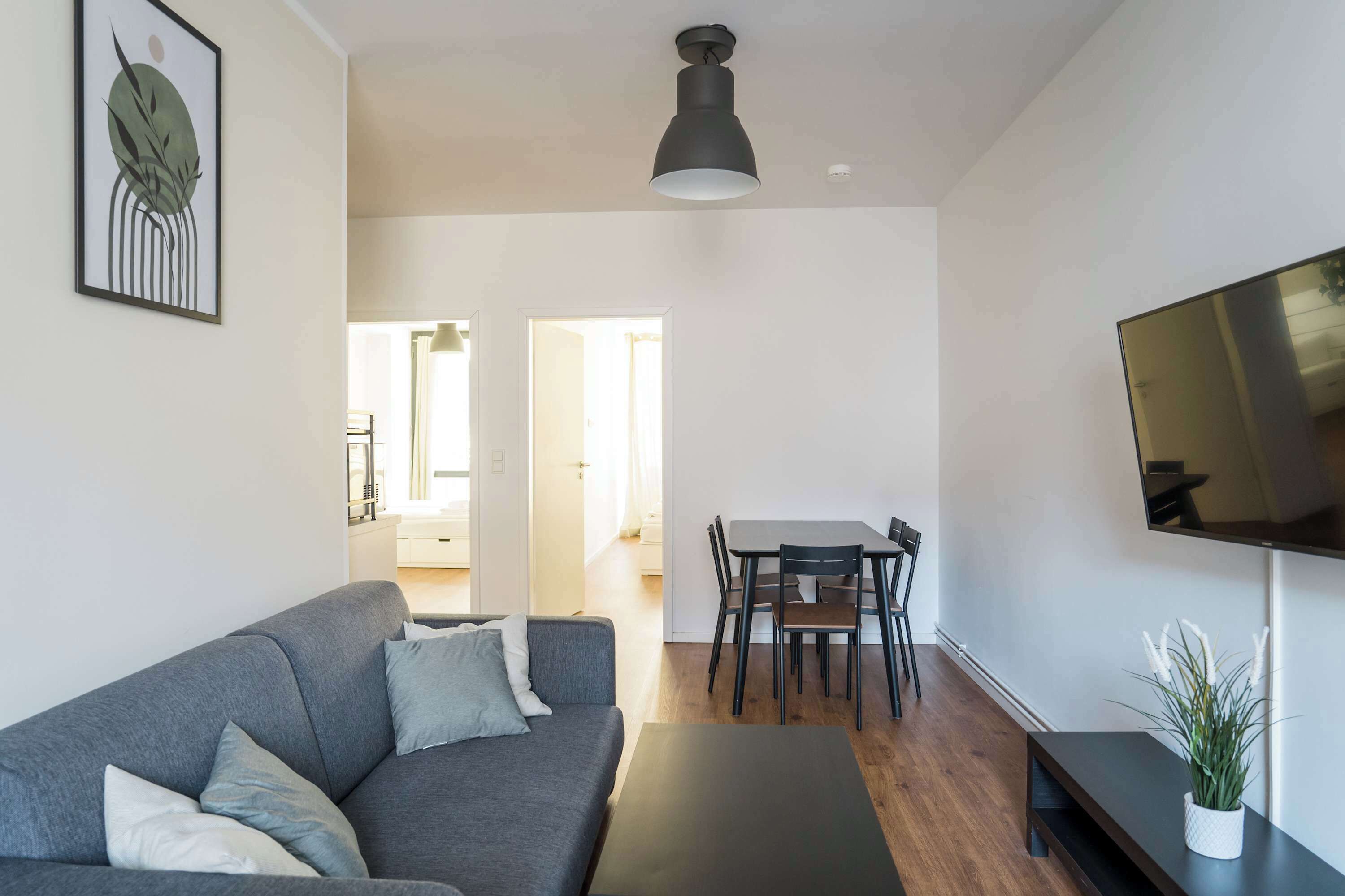 Ein modernes Wohnzimmer mit grauem Sofa, einem schwarzen Couchtisch, einem Essbereich mit Tisch und Stühlen sowie einem an der Wand befestigten Flachbildfernseher.