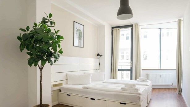 Ein modernes Schlafzimmer mit einem Bett, Nachttischen, einer Topfpflanze und einem Fenster mit Vorhängen.