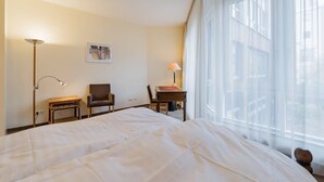 A hotel room with a large bed, a desk, a chair, a lamp, and a window with sheer curtains. - Deluxe Studio in Berlin Mitte (Berlin)
