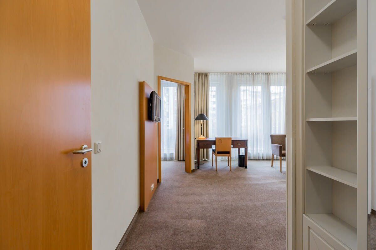 Une chambre d’hôtel avec une porte en bois, un bureau avec une chaise, une télévision et une grande fenêtre avec des rideaux.