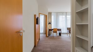 Habitación de hotel con puerta de madera, escritorio con silla, televisor y ventana grande con cortinas.
