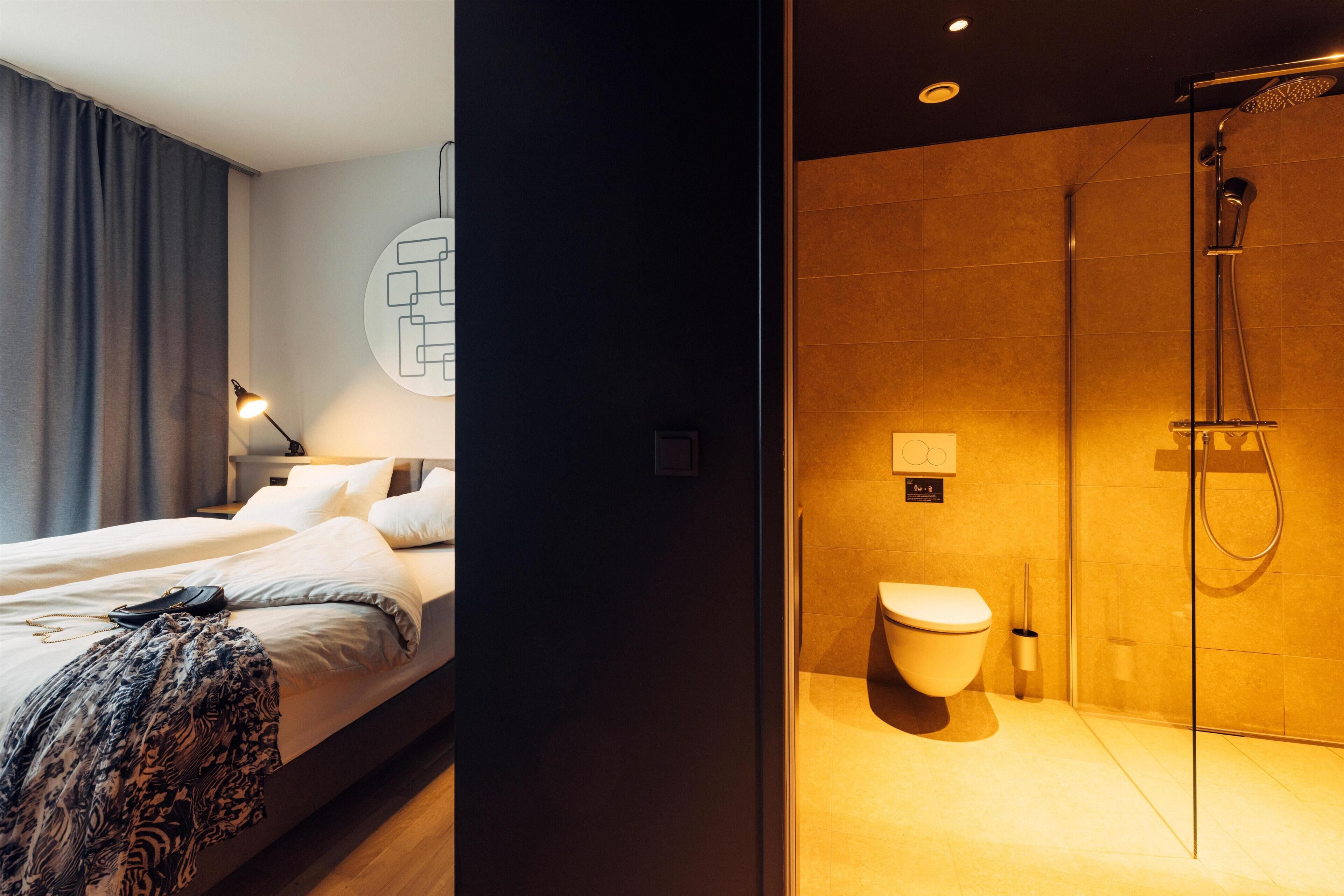 A modern hotel room with a large bed, a bedside table, a lamp, a shower area with a glass door, and a toilet.