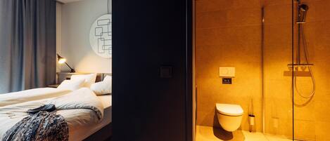 A modern hotel room with a large bed, a bedside table, a lamp, a shower area with a glass door, and a toilet.