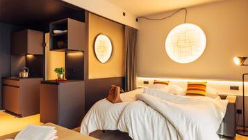 Quarto de hotel moderno com cama, mesas de cabeceira, escrivaninha, cadeira, pequena cozinha e luminária na parede.