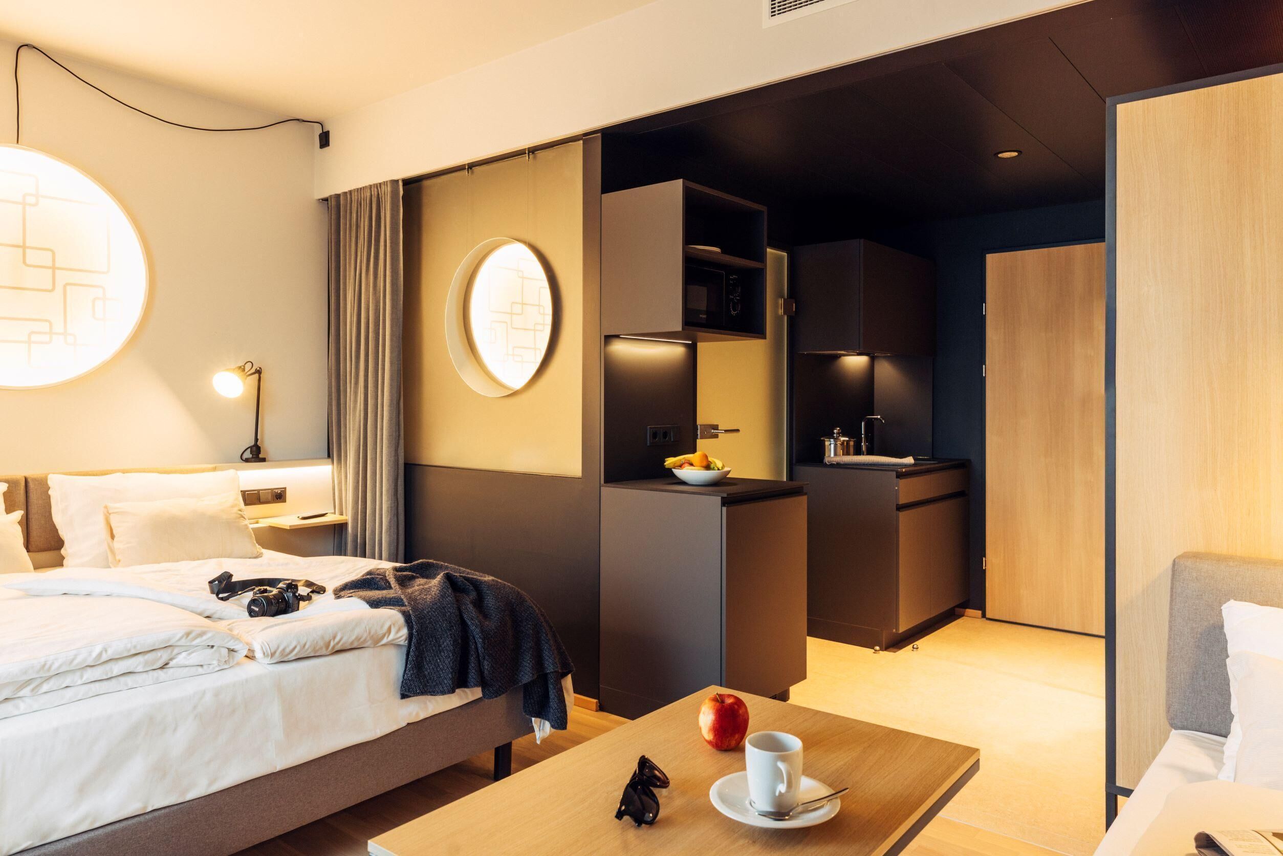 A modern hotel room with a bed, a desk, a small kitchenette, and a wardrobe.
