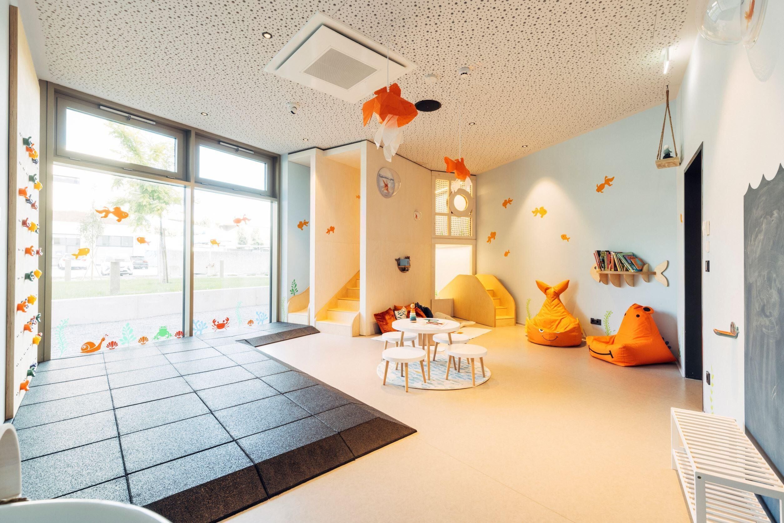 Une pièce lumineuse, dotée de grandes fenêtres, avec une table ronde entourée de chaises, des poufs orange et un berceau blanc.