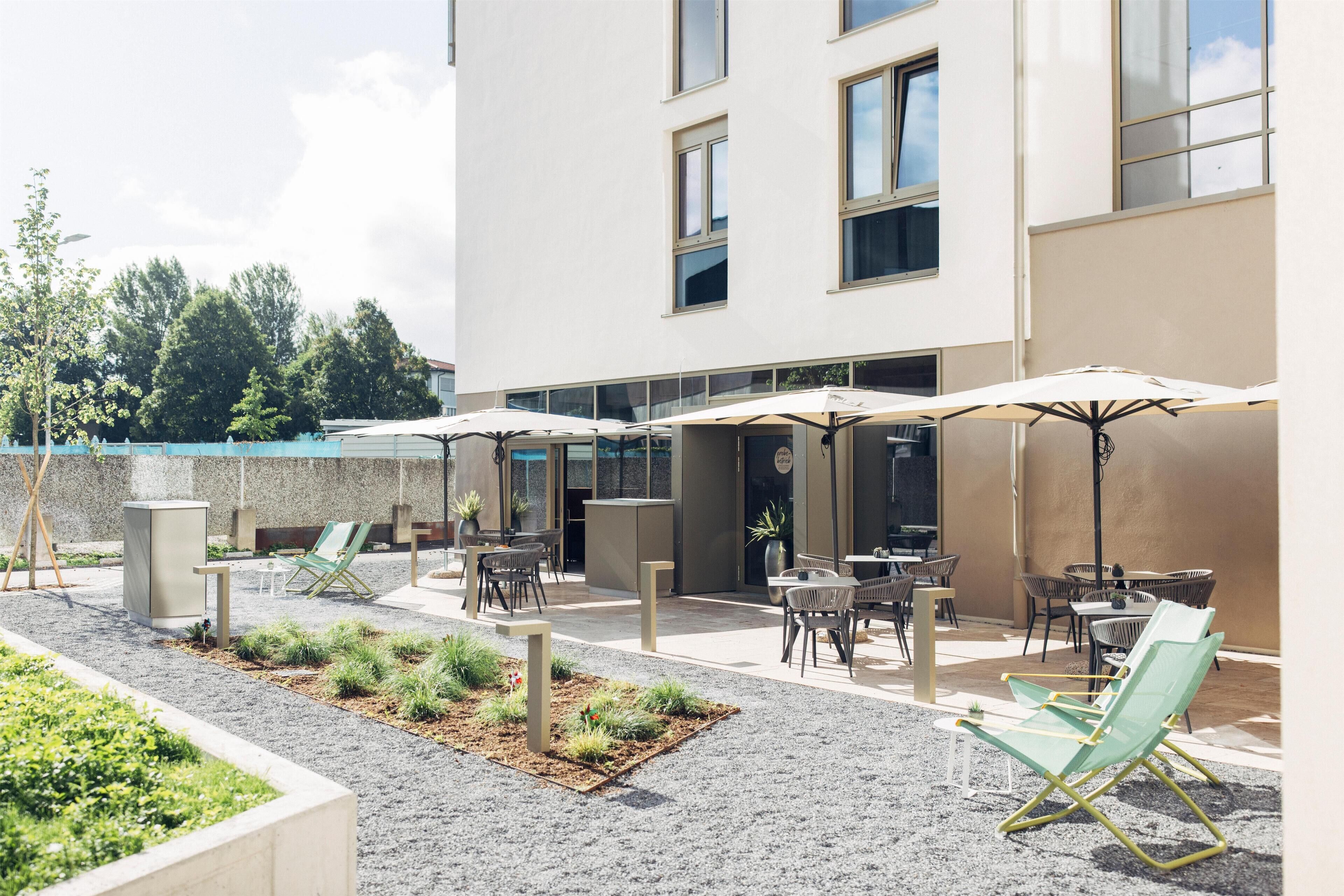 An outdoor seating area with umbrellas and chairs, a building with large windows, and a landscaped garden.