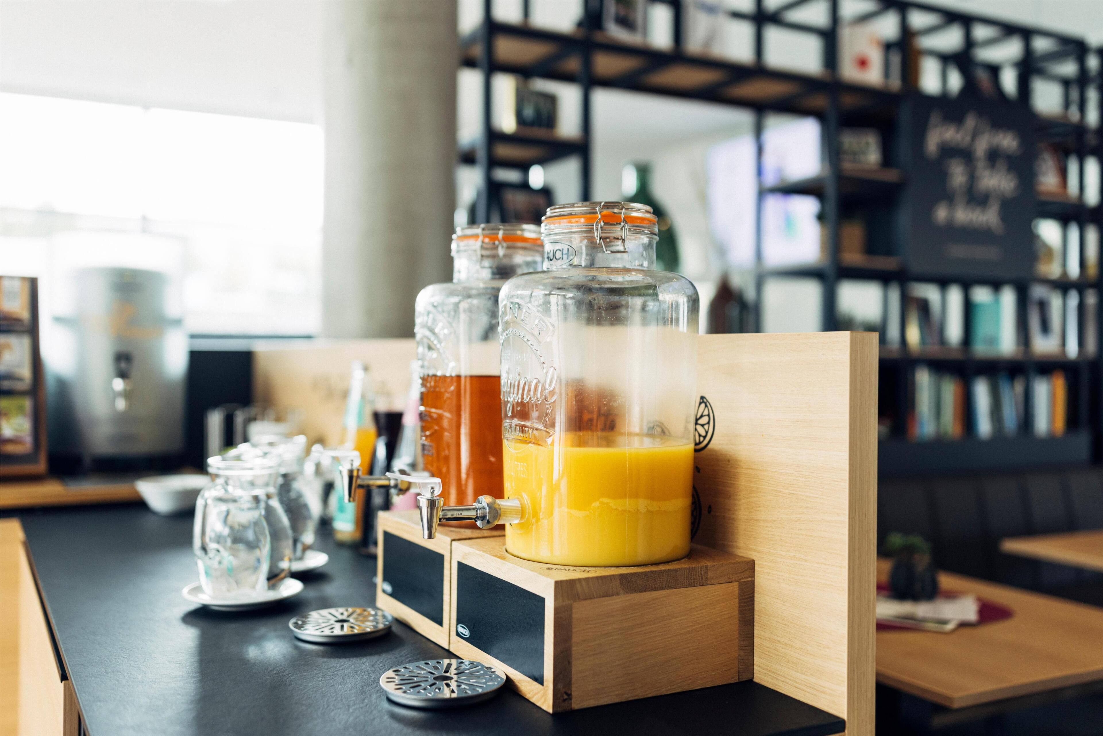 Una zona de bar con mostrador de madera, dispensador de zumo y tarros de vidrio con zumo de naranja.