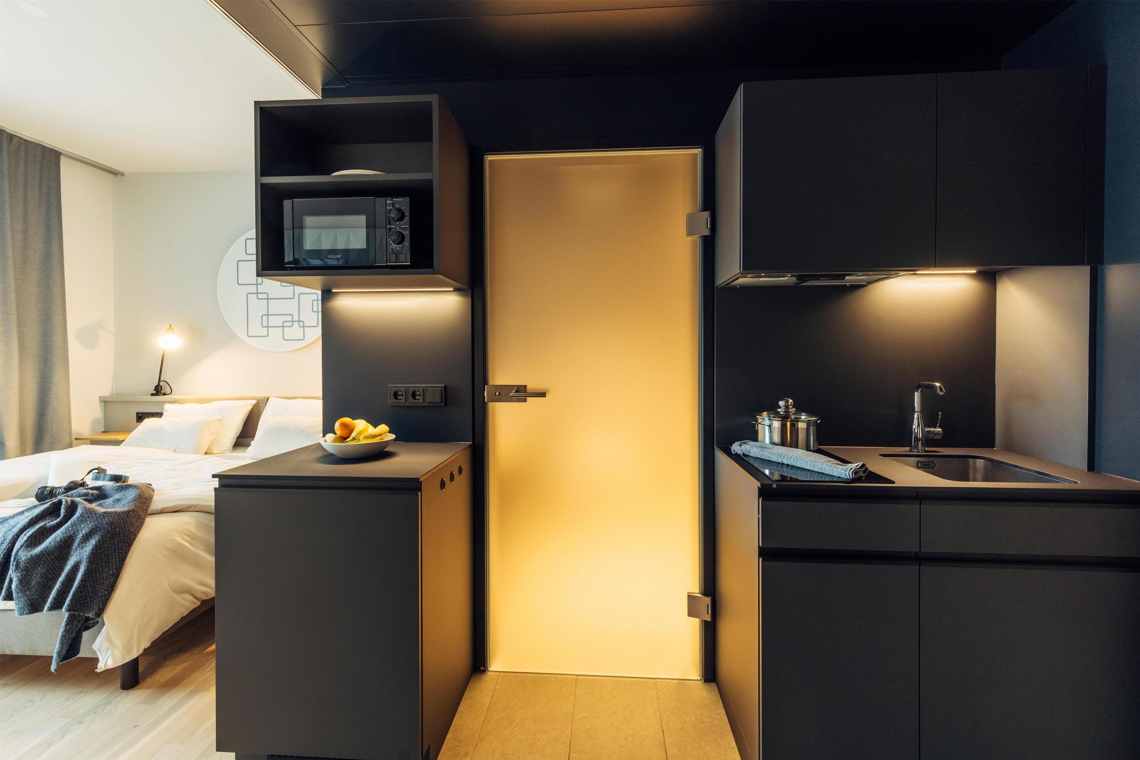 A compact hotel room with a bed, bedside table with fruit, a microwave, a small kitchenette with a sink, and a door leading to another room.