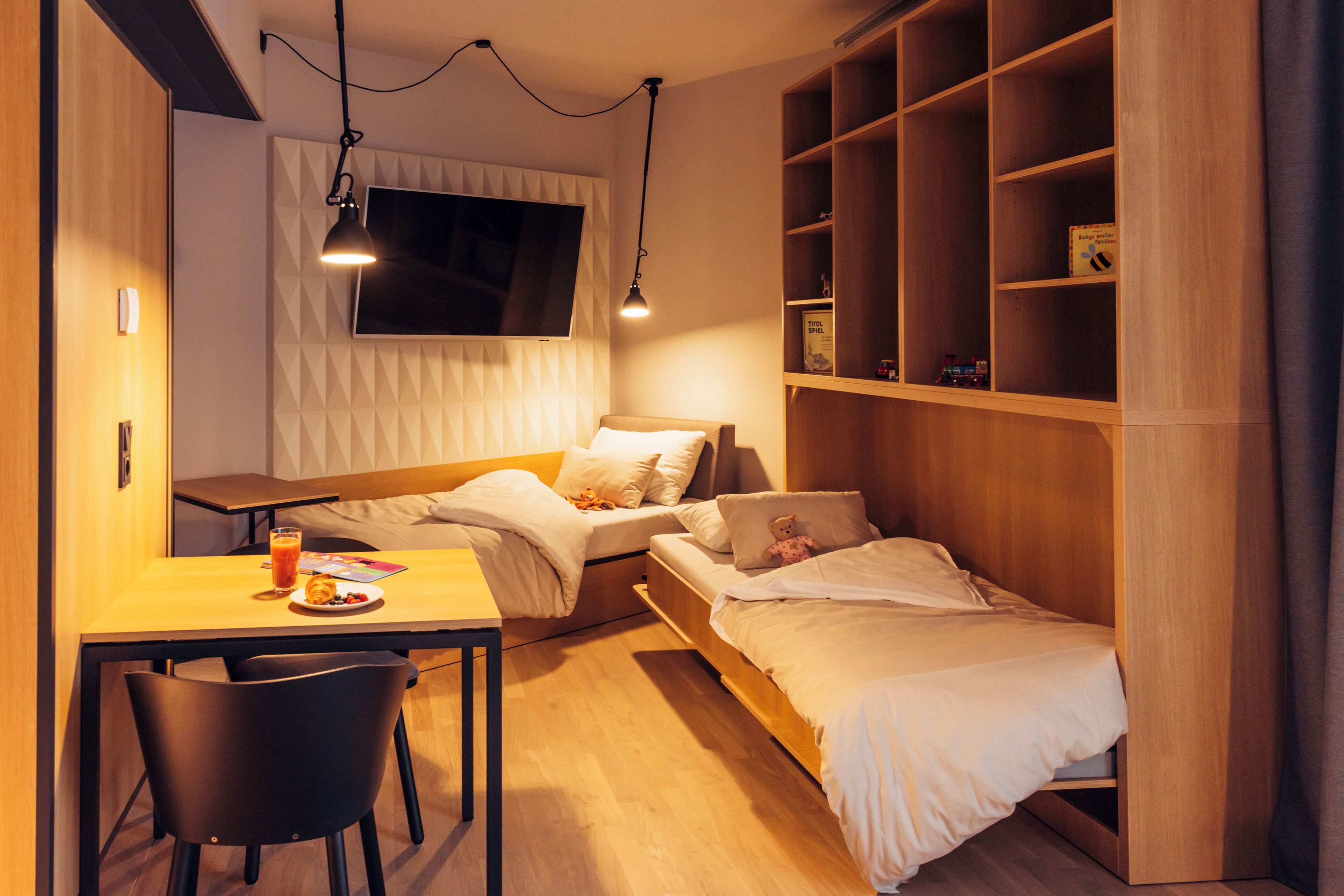 Habitación de hotel compacta con cama, escritorio y silla. Hay un televisor montado en la pared, una estantería con libros y una ventana con persianas.