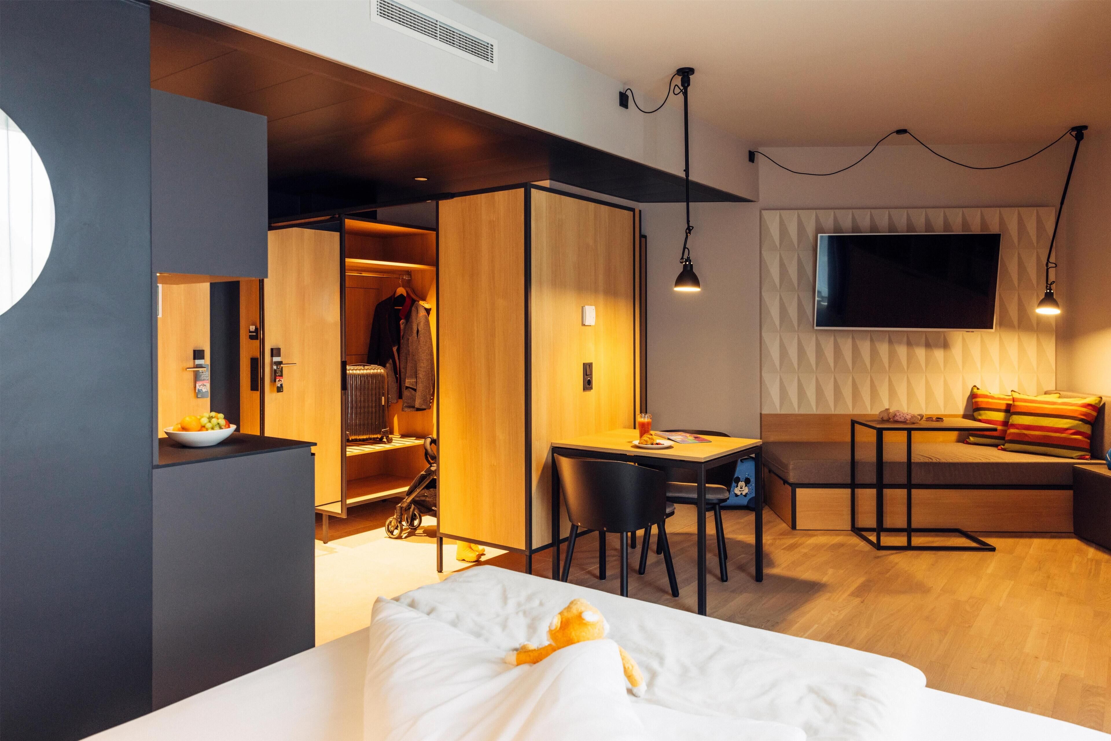 A modern hotel room with a bed, a desk, a chair, a TV, and a wardrobe.