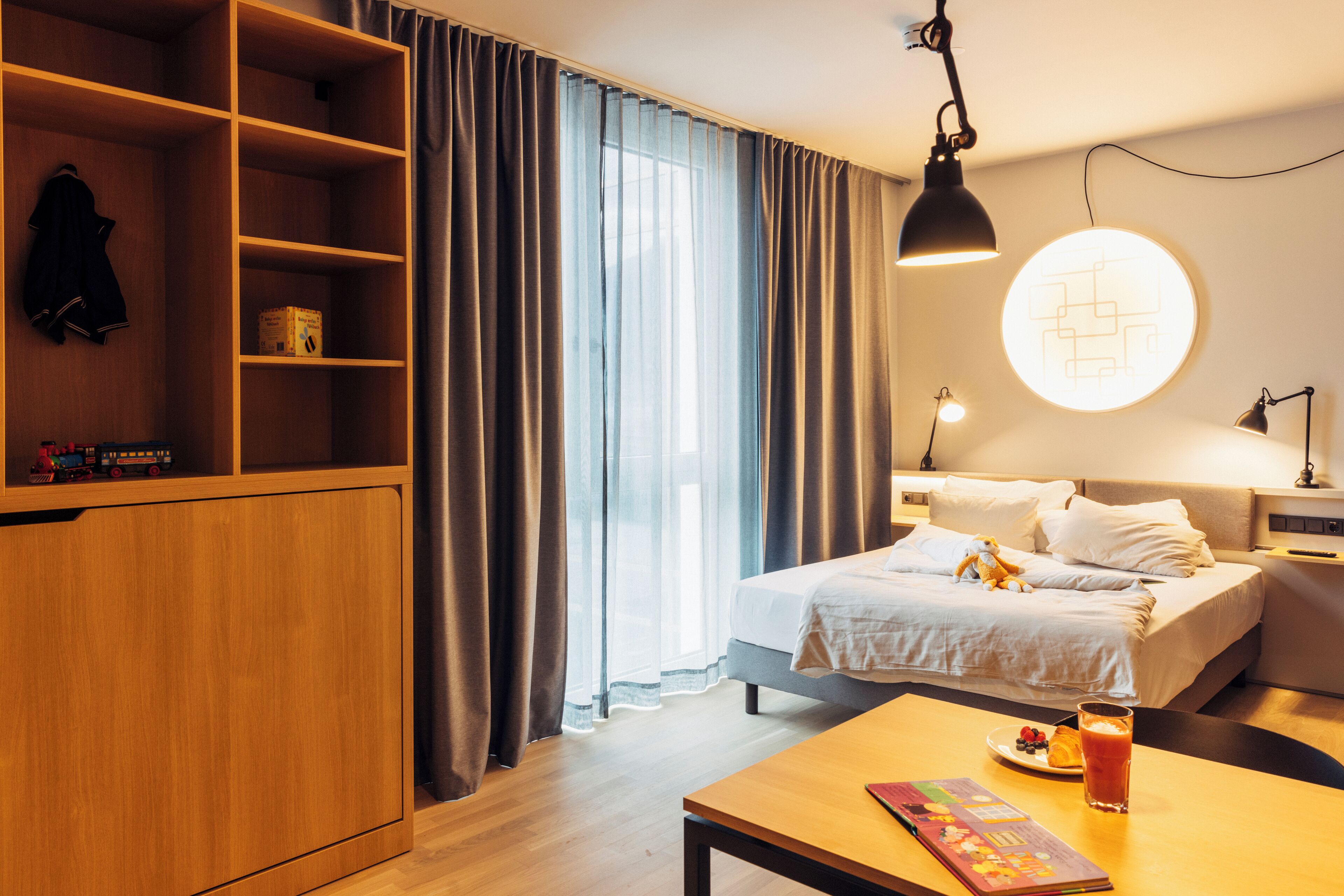Una habitación de hotel con cama, escritorio con una bebida, lámpara y armario.