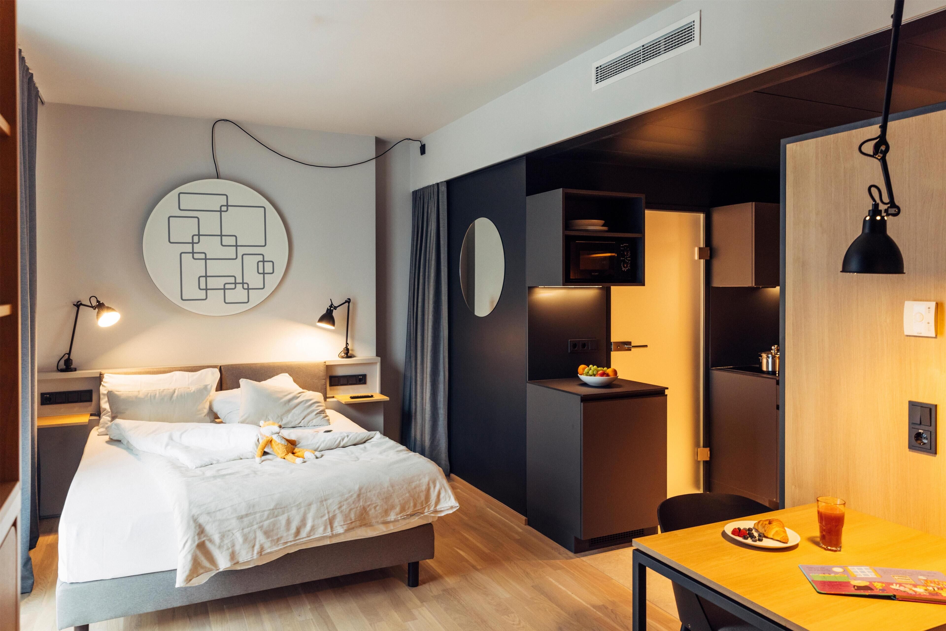 A modern hotel room with a bed, bedside table, and a small kitchenette area.