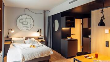 A modern hotel room with a bed, bedside table, and a small kitchenette area.