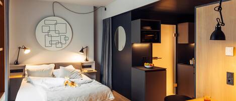 Moderni hotellihuone, jossa on sänky, yöpöytä ja pieni keittonurkkaus.