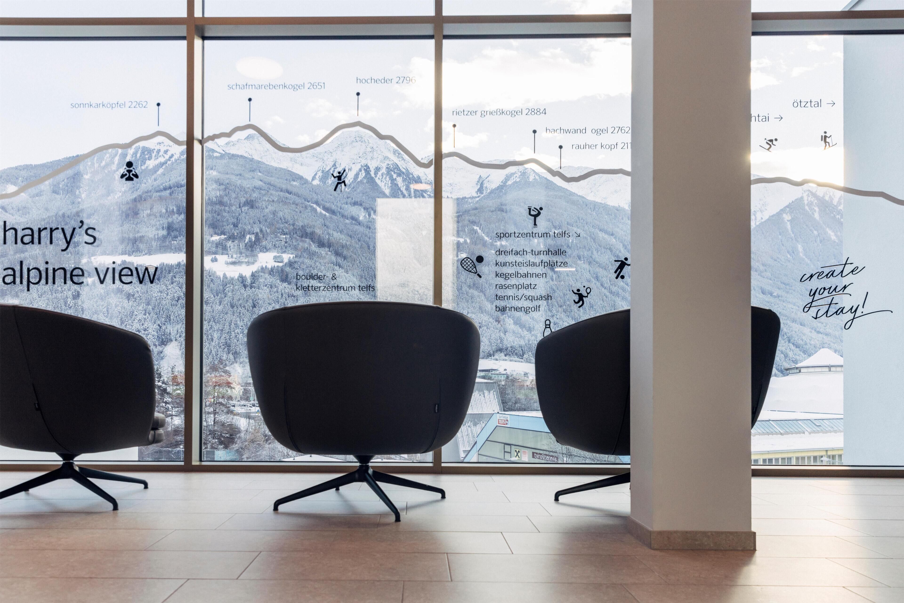 Ein modernes Interieur mit einem großen Fenster, das Blick auf die Berge bietet, zwei schwarzen Stühlen und einer Wand mit Text und Symbolen, die Annehmlichkeiten wie ein Skizentrum, ein Tennisplatz und eine Galerie kennzeichnen.