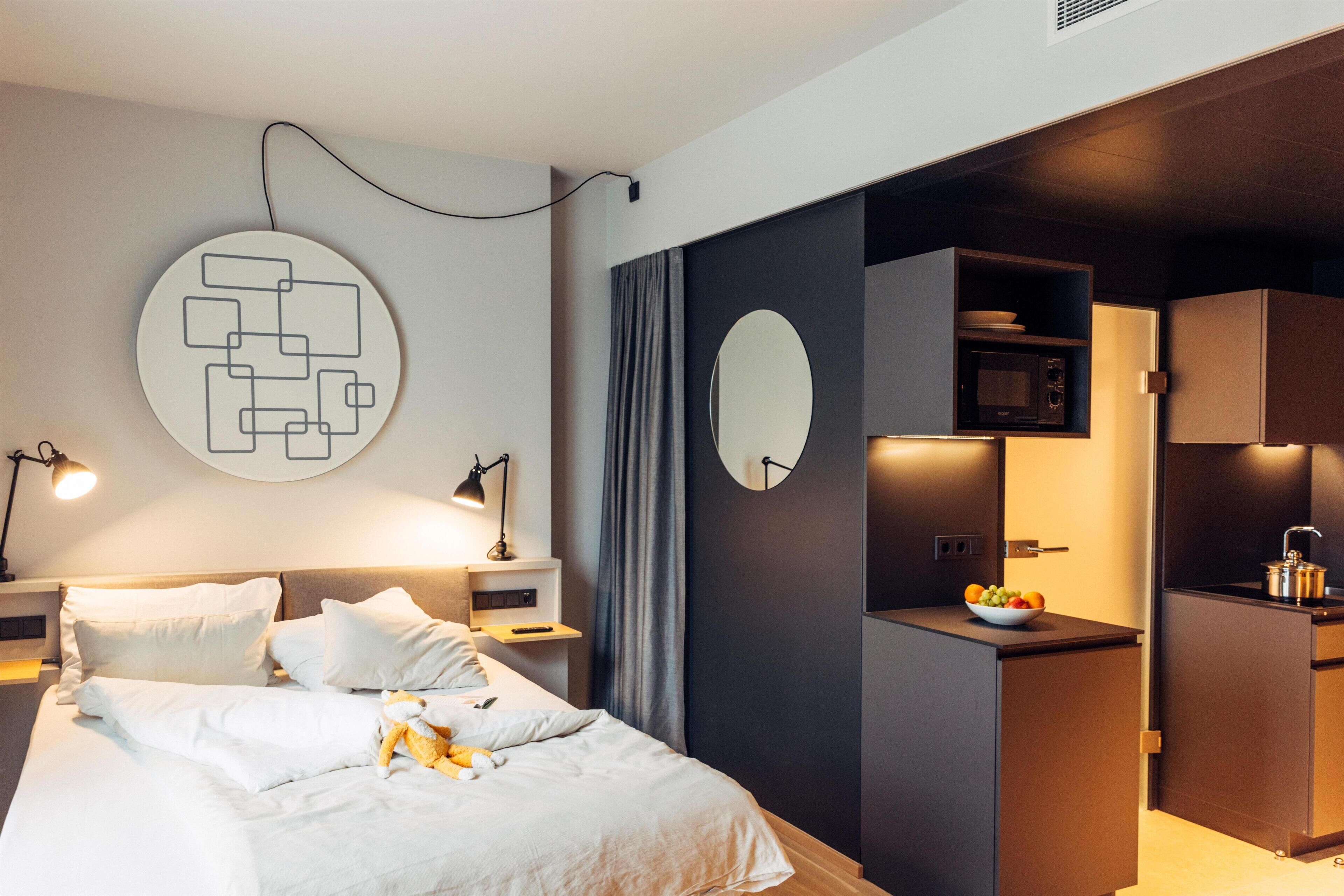 A modern hotel room with a neatly made bed, a wall-mounted mirror, a small kitchenette with a microwave, and a round artwork on the wall.