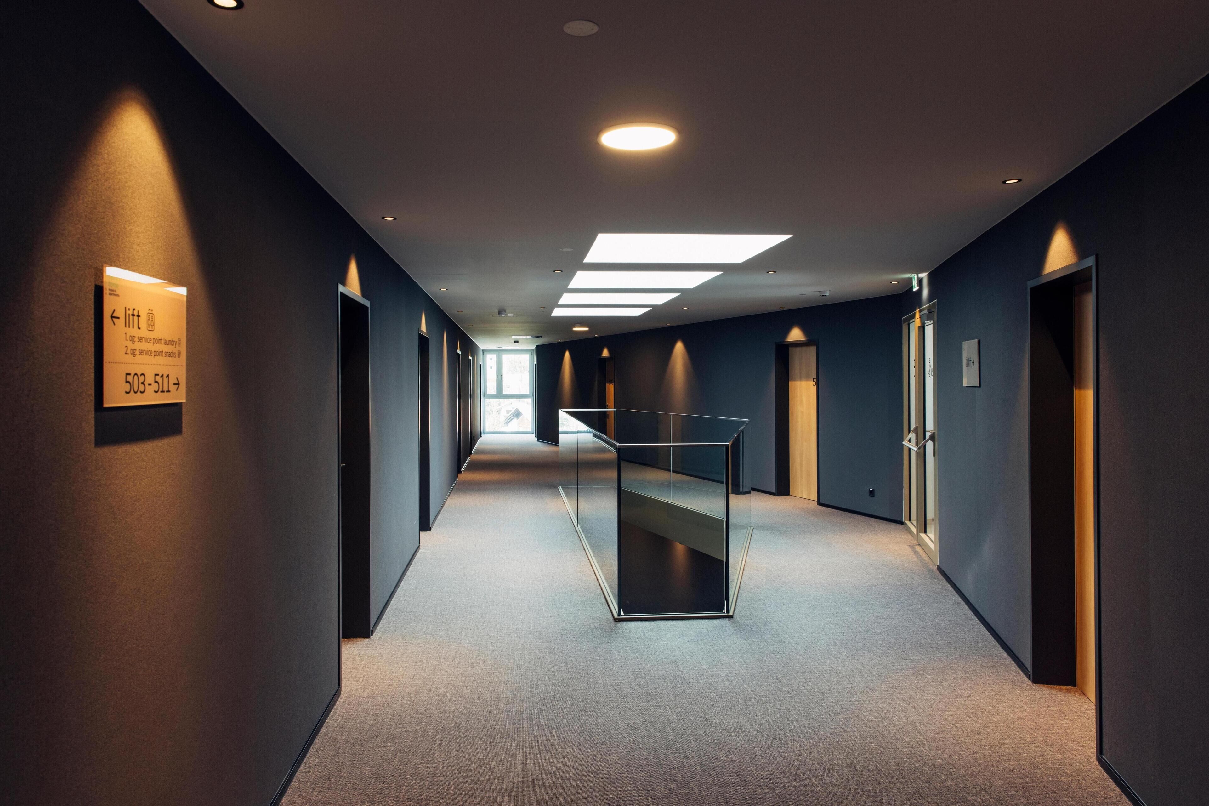 Un couloir d’hôtel moderne, doté d’une cloison en verre, d’un éclairage encastré et d’un panneau indiquant l’emplacement de l’ascenseur.
