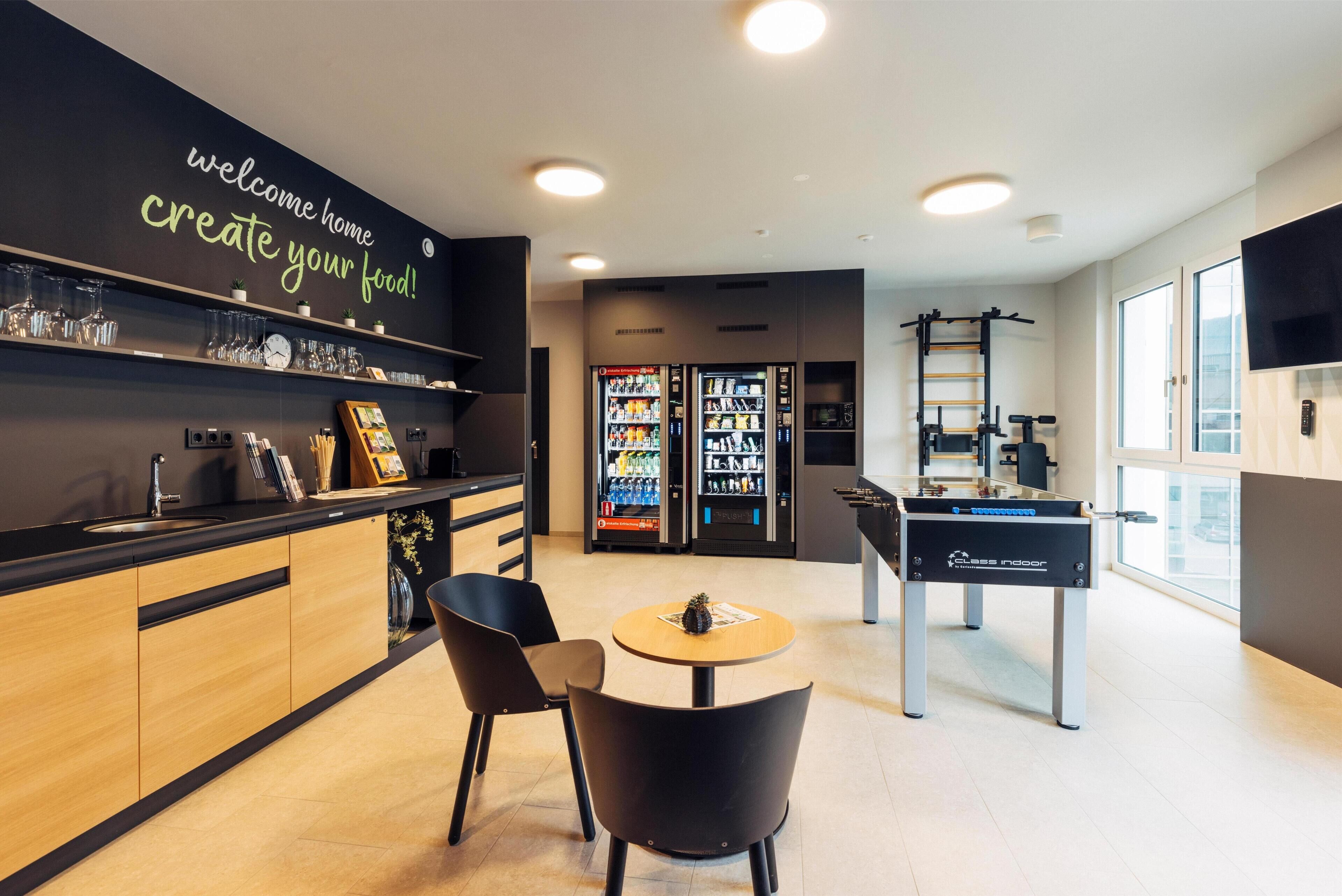 A modern kitchen area with a black countertop, wooden cabinets, and a welcome sign. There are two refrigerators, a coffee machine, and a foosball table.