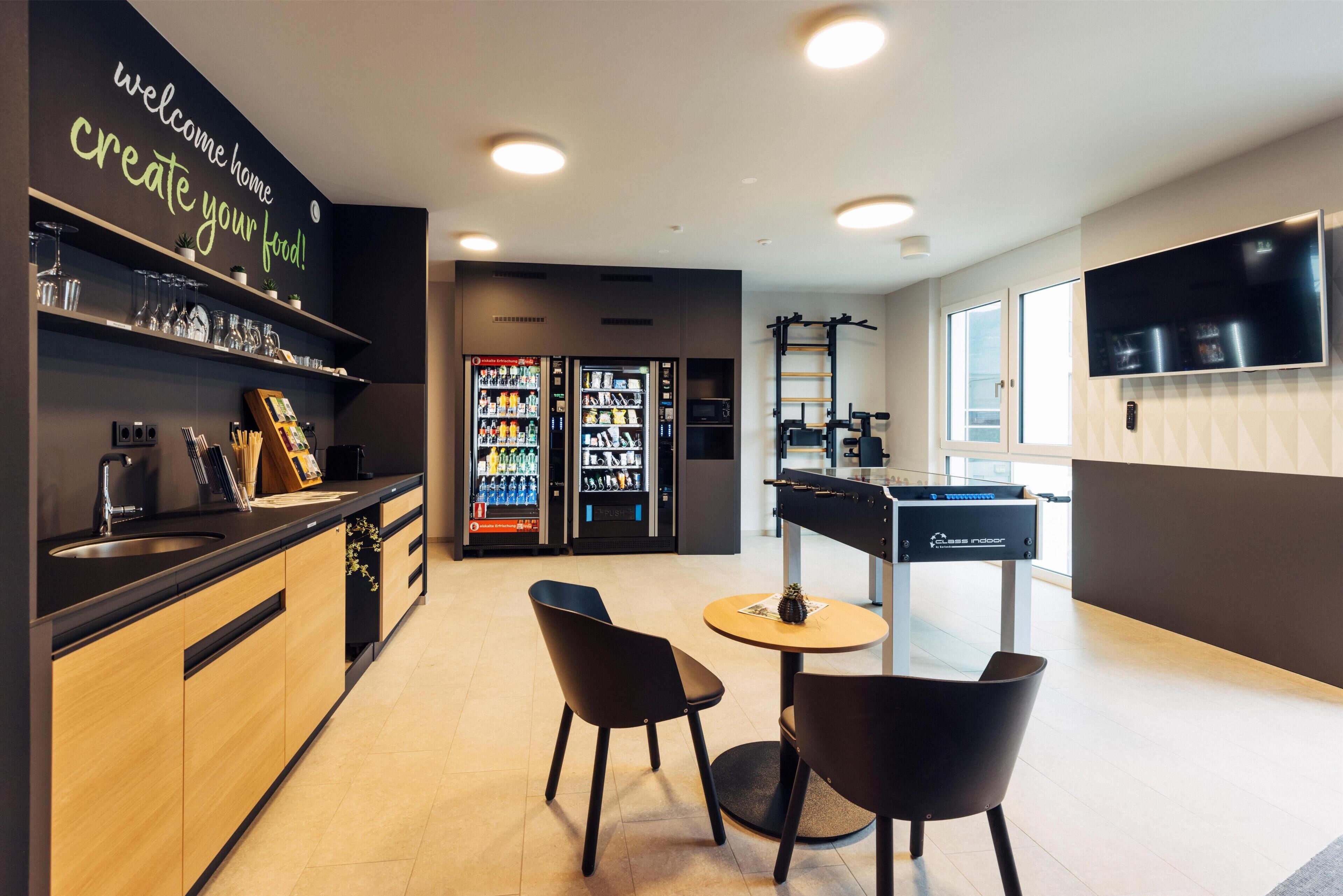 A modern kitchen with a black countertop, wooden cabinets, and a black chair. There are two refrigerators, a coffee machine, and a TV mounted on the wall.