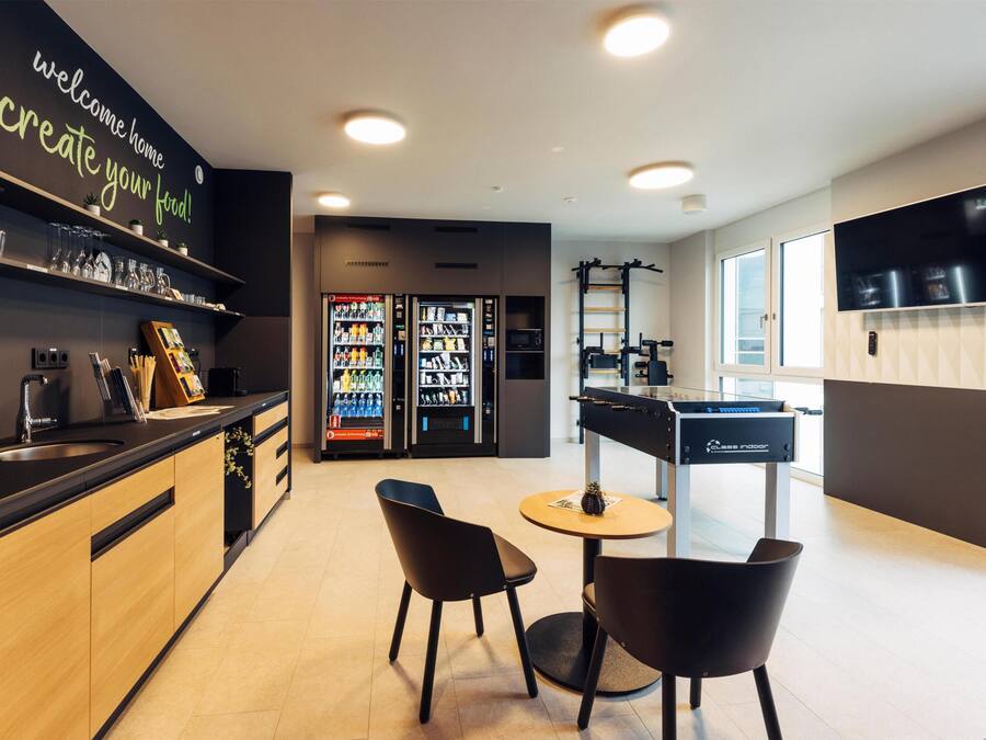A modern kitchen with a black countertop, wooden cabinets, and a black chair. There are two refrigerators, a coffee machine, and a TV mounted on the wall.